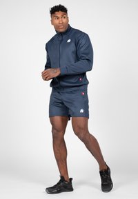 Marineblauwe trainingspak met een ritsjack en shorts, voorzien van een wit logo. Het materiaal is glad en lichtgewicht. Zwarte sportschoenen met een gestructureerd ontwerp.