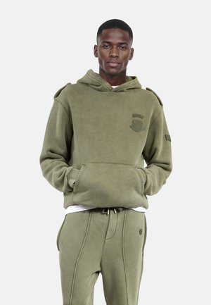 Olijfgroene hoodie met een voorzak, verstelbare trekkoordkap en schouderaccenten. Zacht materiaal met een gestructureerde afwerking.