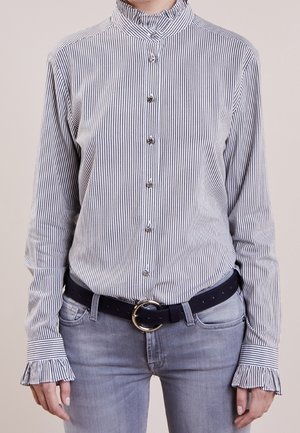 Mujer con una camisa ajustada de rayas verticales y botones, con cuello y puños con volantes, metida dentro de unos vaqueros gris claro con un cinturón negro.