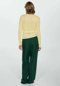 Haut jaune à manches longues côtelé associé à un pantalon large vert à fines rayures. Le fond est neutre, mettant en valeur les détails des vêtements.