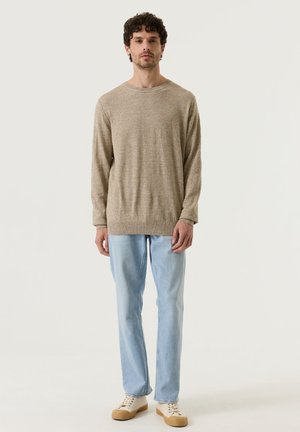 Man met krullend haar, gekleed in een beige trui met lange mouwen, lichtblauwe spijkerbroek en beige sneakers, staand tegen een effen lichte achtergrond.