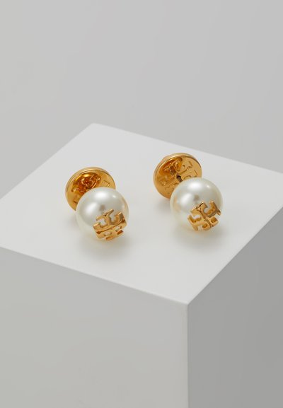 Orecchini a perno in tonalità oro con una grande perla bianca e dettaglio del logo, che evidenziano una texture liscia e una forma rotonda. Design minimalista.