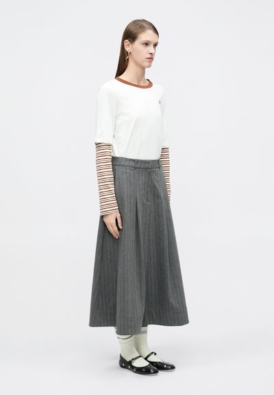 Λευκό μπλουζάκι με καφέ λαιμόκοψη που καλύπτει μια γκρι πλισέ φούστα. Μαύρα παπούτσια με κυκλικά διακοσμητικά και ανοιχτοπράσινες κάλτσες με ρίγες.