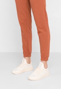 Person som bär rostfärgade joggingbyxor och krämfärgade stickade sneakers, stående på ett ljusgrått golv mot en vit bakgrund.