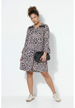 Kvinde med krøllet hår iført en pink og sort leopardmønstret kjole, som holder en sort clutch, iført hvide sneakers, stående mod en hvid væg.