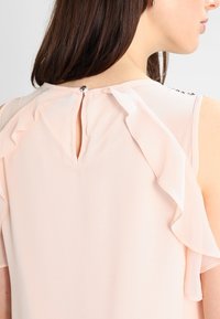 Světlá růžová blůza s volány podél ramen a malým otvorem ve tvaru klíče na zadní straně, zajištěná ozdobným knoflíkem.