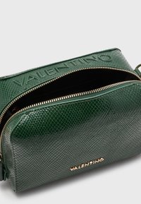 Zelená texturovaná kožená taška se zlatým kováním. Má zipové zapínání a vyražené logo na vnějšku, kompaktní obdélníkový tvar.