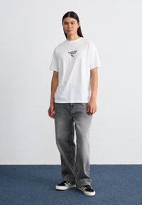 Hvid bomulds t-shirt med sort tekst og logo, parret med grå denim jeans. Modellen har sorte og hvide mønstrede sneakers på.