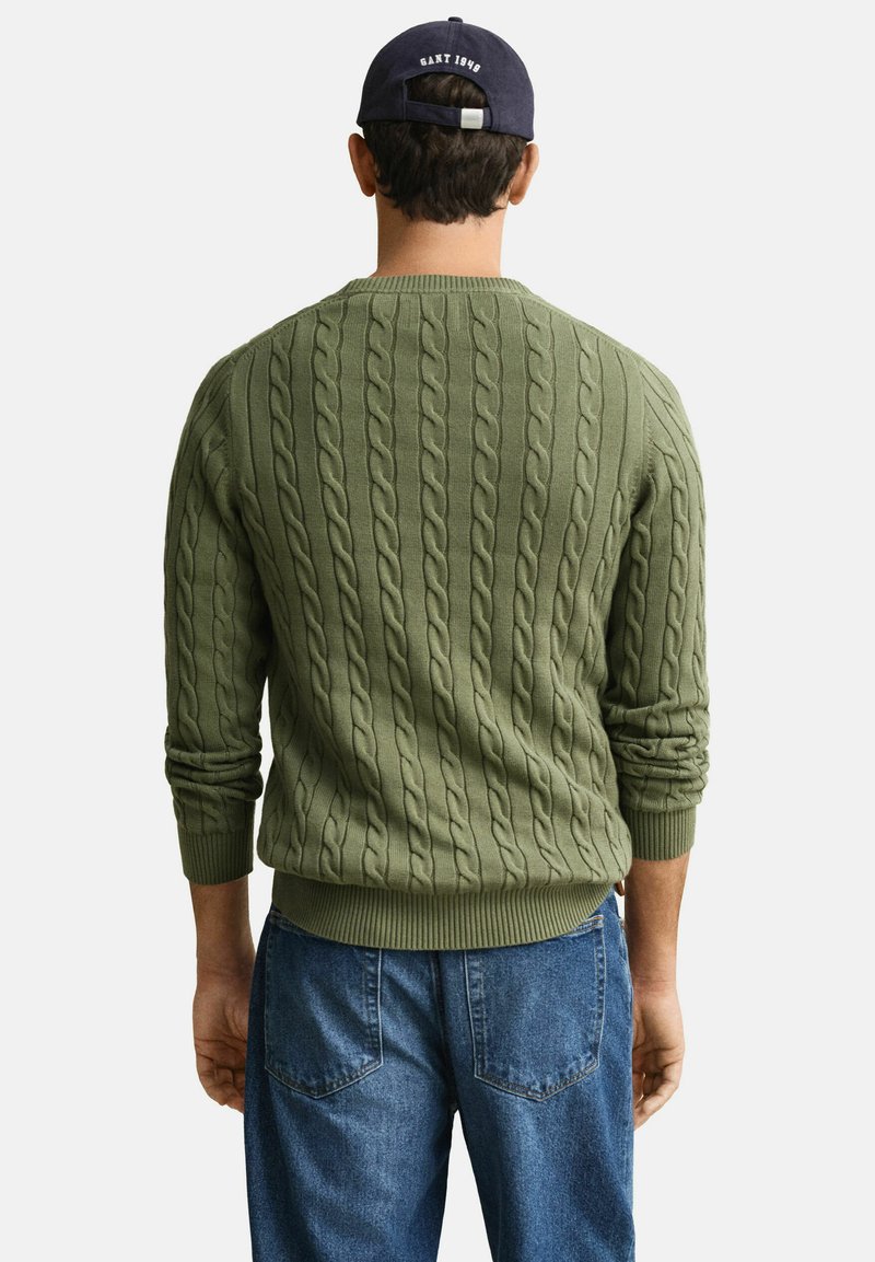 Ein Mann trägt einen olivgrünen, kabelgestrickten Pullover und blaue Jeans. Er steht mit dem Rücken zum Betrachter, trägt eine dunkle Baseballkappe mit der Aufschrift "GANT 1949."