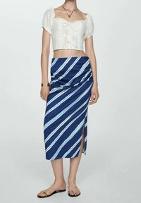 Blouse blanche courte avec manches bouffantes et bordure en dentelle, associée à une jupe midi rayée bleue avec une fente latérale. Sandales simples.