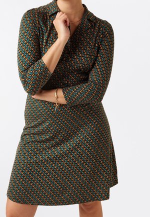 Kobieta ubrana w długą sukienkę w zielono-brązowy wzór, z złotą bransoletką i naszyjnikiem, pozująca z założonymi ramionami i dłonią przy brodzie.