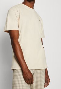 Short-sleeve, light beige cotton t-shirt with a crew neckline and small logo on the left chest. Displays a smooth texture and relaxed fit.