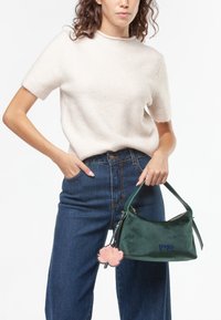 Groene fluwelen handtas met een bloemencharme, voorzien van een ritssluiting en een zachte handgreep, gecombineerd met een crèmekleurige trui met korte mouwen en een blauwe spijkerbroek.