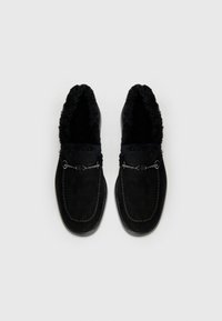 Sorte mokasinslippers laget av semsket skinn, med myk pelsfôr, dekorativ søm, og en metallaccent i midten.