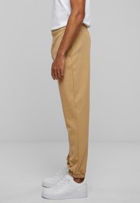Beige Jogginghosen mit elastischem Bund und Bündchen, kombiniert mit weißen Sneakers. Der Stoff wirkt weich und hat eine glatte Textur.