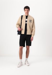 Beige bomber jacket with a logo patch, black t-shirt, and black shorts. White sneakers and a black shoulder bag complete the outfit.