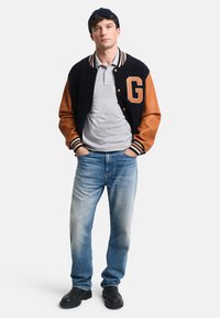 Giacca varsity in lana-acrilico blu navy con maniche in pelle beige, polsini a costine, polo grigia, jeans in denim azzurro chiaro e scarpe nere.