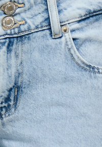 Gros plan sur un jean en denim bleu clair montrant deux boutons argentés, une boucle de ceinture, une poche avant et des coutures détaillées.