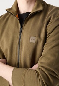 Person med olivgrön zip-up-jacka med BOSS-logotyp på bröstet och svart tröja under, med armarna korsade över kroppen.