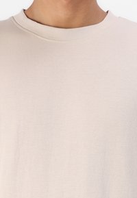 T-shirt à manches courtes beige clair avec un col rond. Le tissu semble doux et lisse, présentant un design uni sans motifs ni accents.