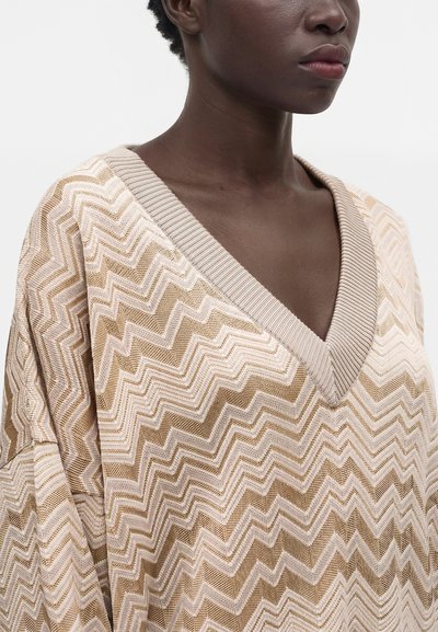 Personne à la peau foncée portant un pull à col en V beige et marron à motif zigzag sur un fond blanc uni.