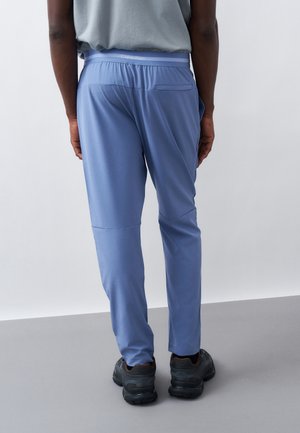 Άτομο που φοράει ανοιχογάλαζο casual παντελόνι με ελαστική μέση και μαύρα αθλητικά παπούτσια, στέκεται μπροστά σε έναν απλό άσπρο τοίχο και γκρι πάτωμα.