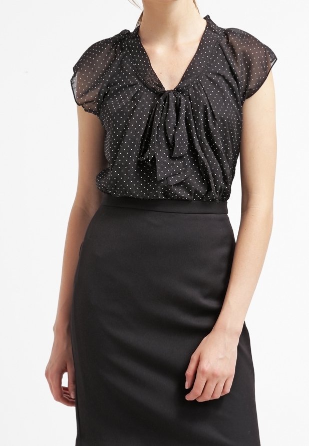 Femme portant un chemisier noir transparent à pois avec un nœud au col et une jupe crayon noire taille haute sur fond blanc.