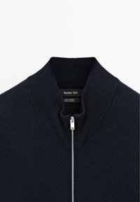 Dunkelblaues Kaschmirpullover mit silbernem Reißverschluss und hochgeripptem Kragen, Etikett liest "Massimo Dutti", hergestellt in Kambodscha.