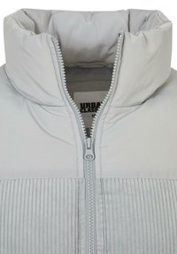 Chaqueta acolchada de color gris claro con un cuello alto, cierre frontal con costuras de acento y textura acanalada en la sección inferior.