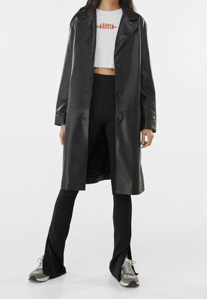 Vrouw draagt een lange zwarte leren jas over een wit cropped top met de tekst "ANGELS", zwarte split-zoom broek en grijze sneakers.