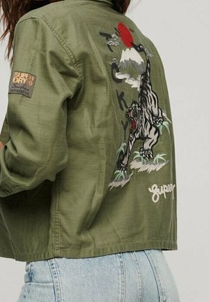 Chaqueta verde oliva con tigre bordado, montaña, sol y texto "Tokyo" en la espalda, usada con vaqueros azul claro.