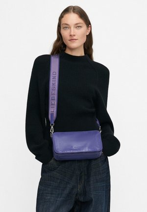 Donna con lunghi capelli castani che indossa un maglione nero e jeans scuri, mentre porta una borsa a tracolla viola con una larga tracolla sulla spalla.
