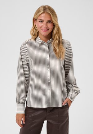 Femme blonde portant une chemise à rayures verticales blanches et noires boutonnée et un pantalon marron foncé, souriante, une main dans la poche.