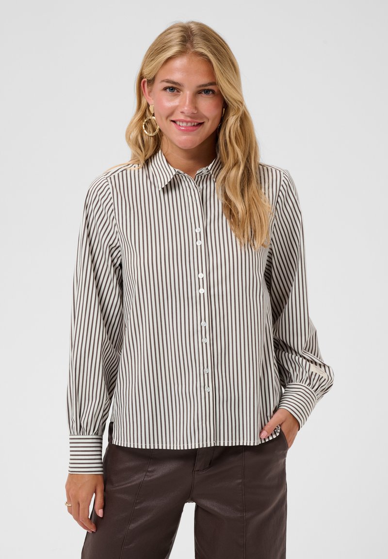 Femme blonde portant une chemise à rayures verticales blanches et noires boutonnée et un pantalon marron foncé, souriante, une main dans la poche.