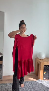 Femme avec les cheveux en chignon tenant une robe rouge à manches longues sur un cintre dans une pièce lumineuse avec des meubles en bois et un sol moquetté.