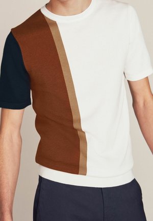 Homme portant un pull à manches courtes à blocs de couleurs blanc, marron, beige et bleu marine, associé à un pantalon bleu foncé.