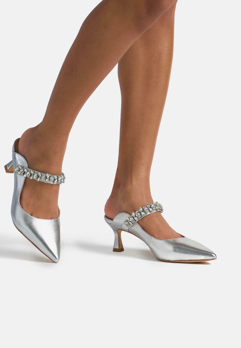 Mules plateadas con punta afilada, que cuentan con una tira decorativa de rhinestones y un tacón bajo y ancho. Textura metálica suave.