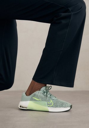 Stopa w jasnozielonym meshowym sneakersie z neonowymi akcentami, ustawiona w pół kroku na gładkiej podłodze, ubrana w czarne spodnie.