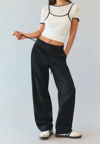 Vrouw in zwarte wijde broek met touwtjes, witte crop top met zwarte rand en witte sneakers, staand tegen een lichte achtergrond.