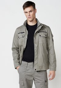 Svetlošedá bunda na zips s viacerými vreckami, vrátane dvoch vreciek na hrudi a jedného vrecka na hrudi so zipsom. Vyrobená z ľahkého materiálu.