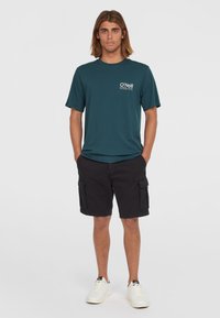 Tyrkysové bavlnené tričko s malým bielym logom na ľavej hrudi, skombinované s čiernymi cargo šortkami a bielymi teniskami. Uvoľnený outfit s voľným strihom.