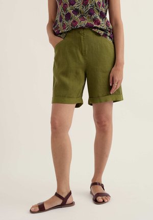 Vrouw draagt olijfgroene shorts met opgerolde zoom, bloemenmouwloos topje en bruine platte sandalen, staand met één hand in de zak.