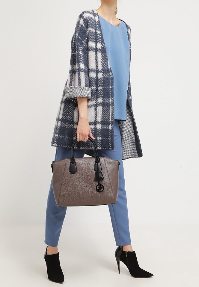 Femme portant une tenue bleue et un manteau à carreaux tenant un grand sac taupe avec des poignées noires, portant des bottines noires à talons hauts.