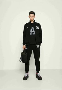 Νεαρός άνδρας σε μαύρη φόρμα New York Yankees, μαύρο καπέλο, κρατώντας μια μικρή μαύρη τσάντα, που στέκεται μπροστά σε απλό ανοιχτό φόντο.