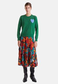 Suéter verde con un parche de corazón azul, combinado con una falda de capas multicolor que presenta patrones abstractos. Botas negras hasta la rodilla completan el look.