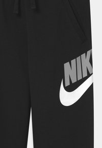 Calças de treino pretas com cintura elástica, apresentando um grande Swoosh da Nike em branco e texto "NIKE" em cinza na perna esquerda. Tecido macio.