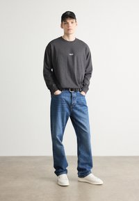 Cămașă gri cu mânecă lungă, dotată cu un mic logo, blugi albaștri din denim, adidași albi și o șapcă neagră cu text. Ținută casual, cu o croială lejeră.