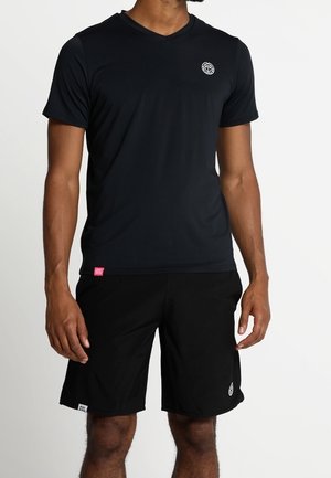 Mann trägt ein einfarbiges schwarzes V-Ausschnitt-T-Shirt und schwarze Shorts mit kleinen weißen Logos auf der Brust und dem Bein vor einfarbigem Hintergrund.
