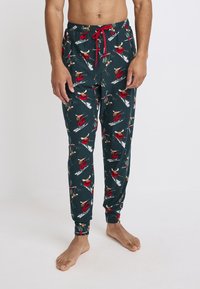 Groene pyjamabroek van zacht materiaal, met feestelijke patronen van skiërs en elanden in het rood, voorzien van een elastische tailleband en een rode aansnoerkoord.