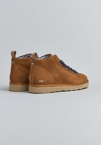 Un paio di stivali ankle in suede marrone con lacci blu navy, suole beige, fodera in pelliccia interna e linguette sul tallone, su uno sfondo grigio.